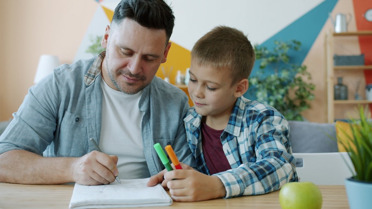 Caring father creative man is drawing pictures while kid is watching sitting at desk in house enjoying time together. Family activities and lifestyle concept.